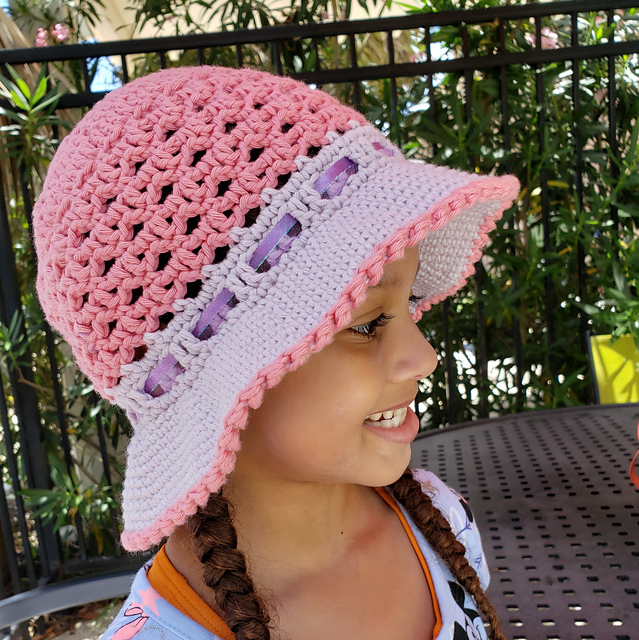 A pink crocheted cotton sunhat with purple brim and a purple ribbon detail, modelled on a smiling child with brown braided pigtails
