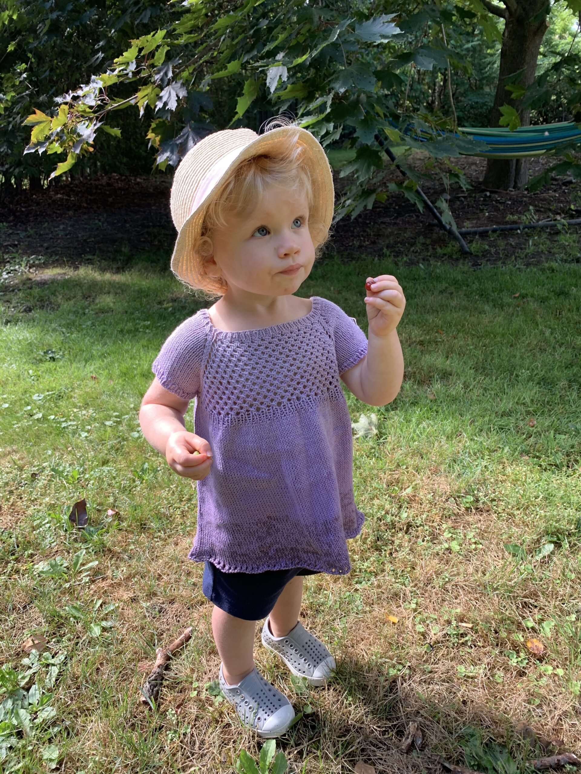 A blonde toddler in a sun hat, standing in a garden picking berries, modelling a lilac ombre babydoll tee with lace bodice and eyelet hem.
