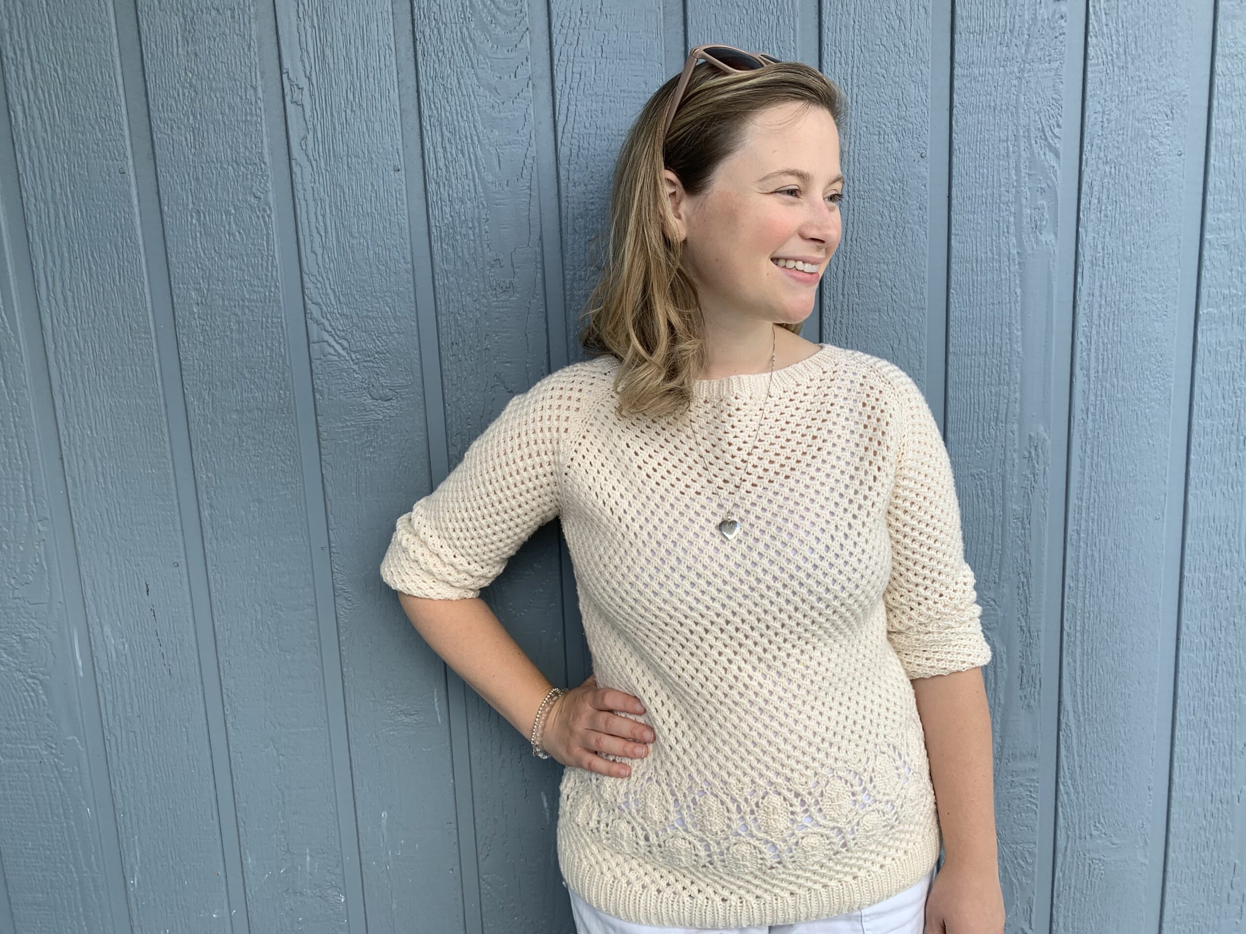 A cream pullover raglan in DK weight yarn in an allover mesh stitch with sand dollar lace motif across the hips, modelled on a woman with 1 inch positive ease