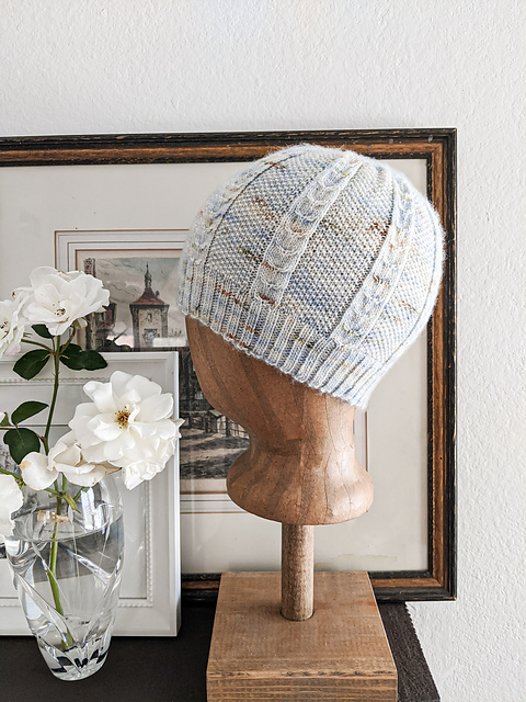 A fitted knit hat with columns of cables between sections of seed stitch, with a ribbed brim, knit in pale grey, pale blue and tan variegated yarn. The hat is displayed on a head form with a vase of white roses and some framed art.