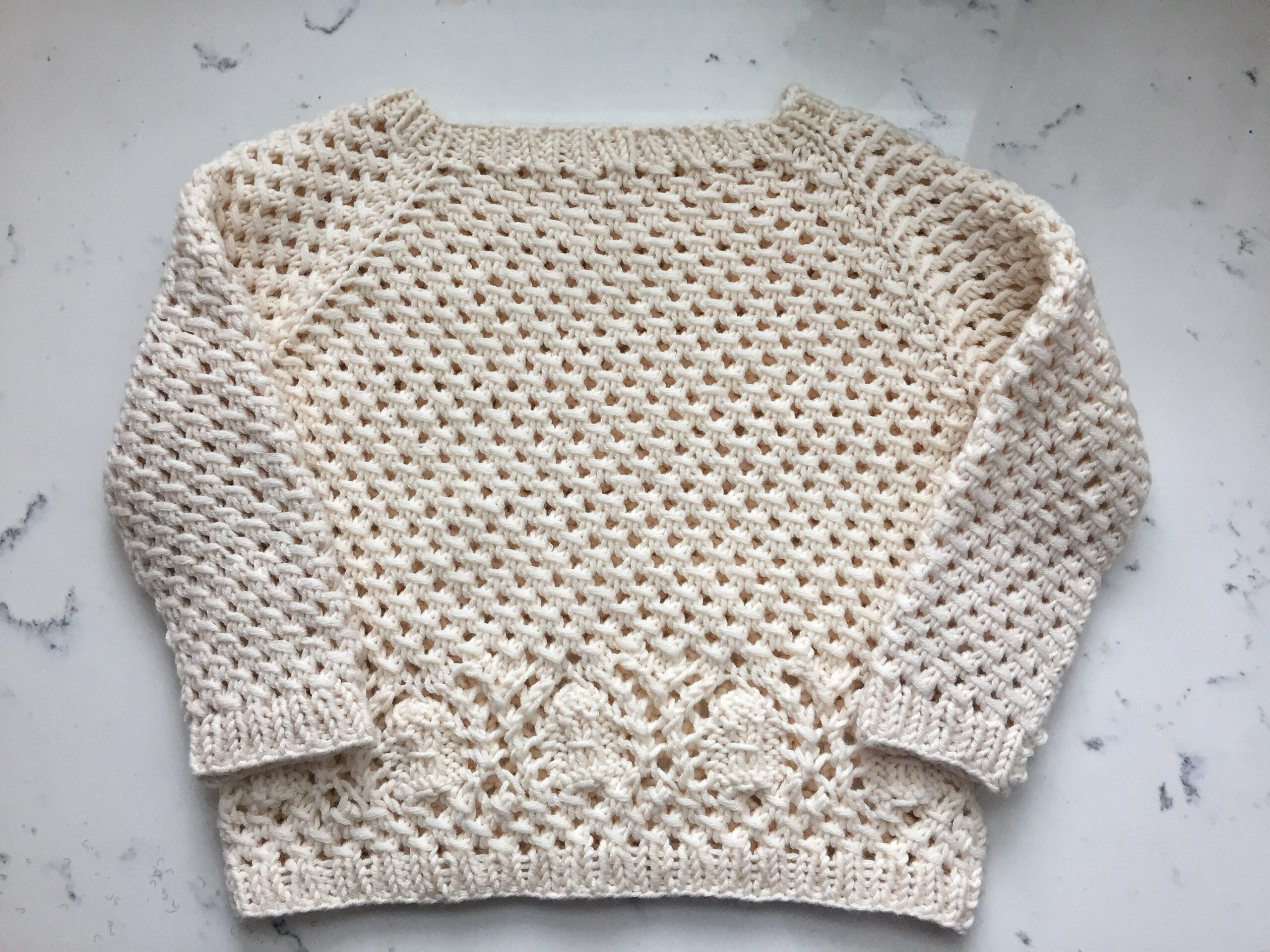 A cream pullover raglan in DK weight yarn in an allover mesh stitch with sand dollar lace motif across the hips