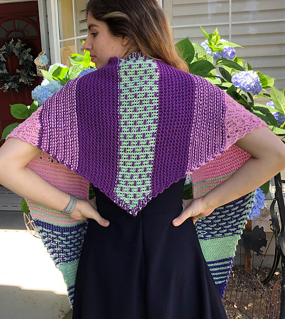 A young woman in a black dress, facing away, modelling a pink, puple, and mint triangle shawl with stripes, eyelets, and colourwork details.