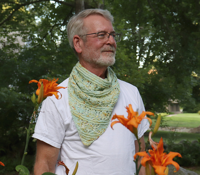 A grey-haired man modelling a pale green garter and lace bandana cowl, standing in a garden of orange lilies.