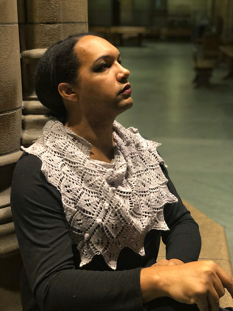Calista, a black trans woman, looks up and to the right with a confident expression on her face. She is seated with her back straight and her hands rested on her lap. She has The Audacity Shawl, a gray lace shawl with a bobble detail, wrapped around her neck and shoulders.
