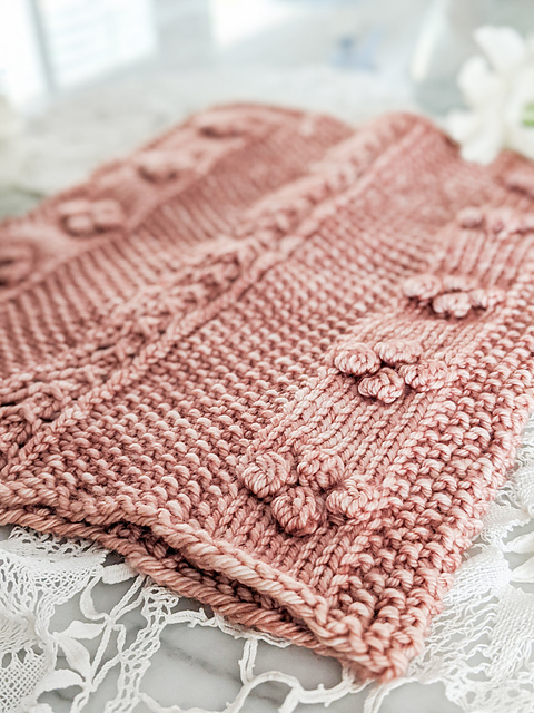 Close up of a seed stitch and bobble cowl, laid flat, knit in tonal rose coloured yarn.