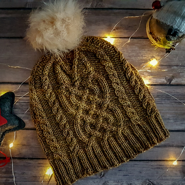 A winter hat with celtic knot cables and ribbed brim, knit in tonal gold dk weight yarn, with a fur pom pom