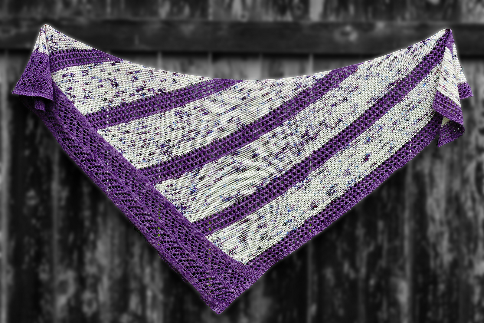 A triangle shawl with sections of white and purple speckled garter, separated by purple strips of eyelet lace.