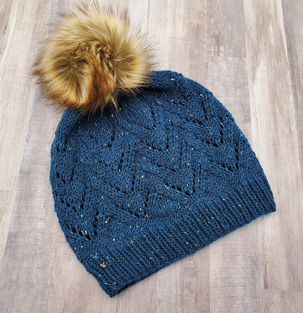 A teal tweed toque with lace detail and a brown fur pom pom
