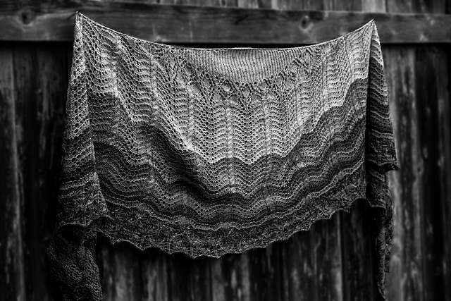 A black and white photo of a lace crescent shawl draped on a fence.