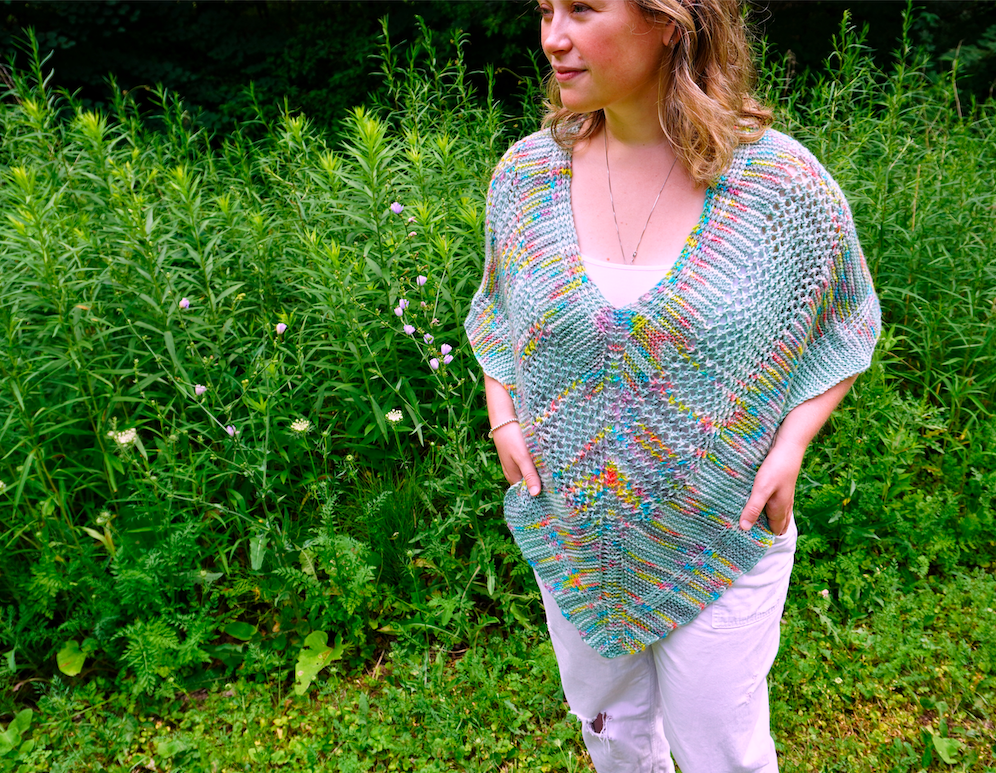 A woman with wavy blonde hair standing in a park, models a grey and rainbow v-shaped poncho