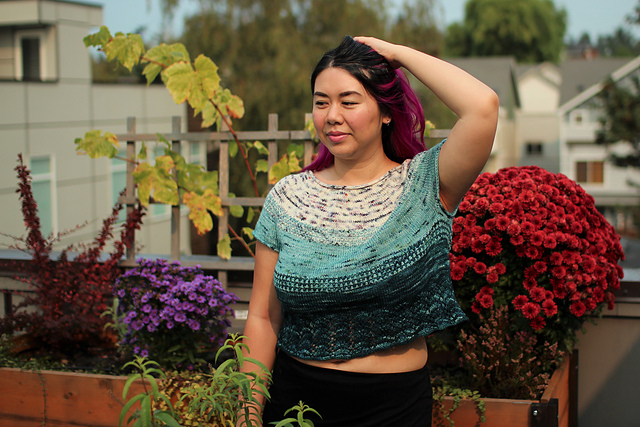 A woman with black and pink hair models a cropped lace tee that fades from off white with black speckles at the yoke, to light teal with black speckles, to tonal deep teal at the hem